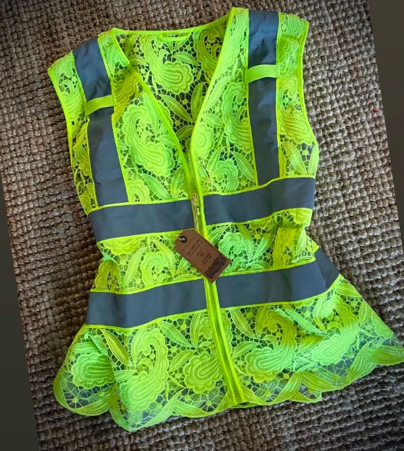A Safety Yellow lace vest laid flat on a jute mat, grey reflective bands across the middle and over the shoulders, with a brown swing tag on the zip.