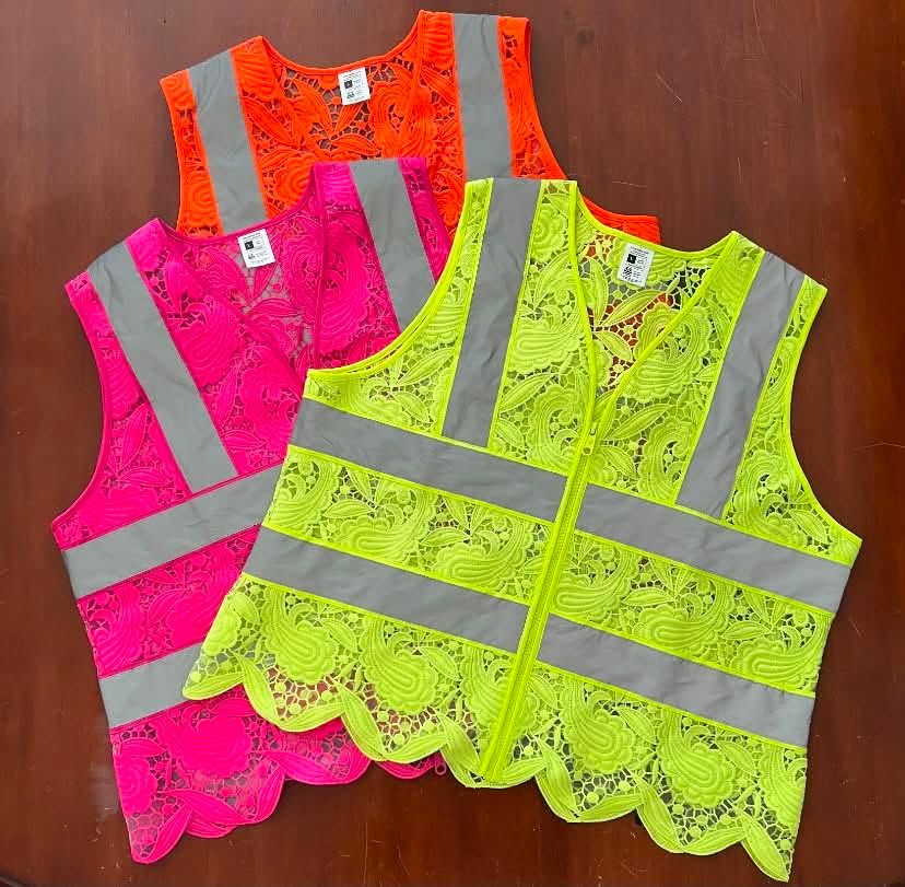 Three lace vests fanned out on a wooden table: orange at the back, pink on the left, Safety Yellow in front, each with grey reflective bands and a scalloped hem.